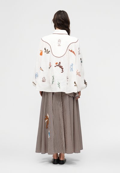 Γυναίκα που φοράει φαρδιά λευκή μπλούζα με πολύχρωμη αφηρημένη κεντητική και μακριά φούστα με καφέ ρίγες, απεικονίζεται από πίσω, όρθια μπροστά σε απλό φόντο.