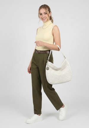 Joven mujer sonriendo, lleva un jersey de cuello vuelto amarillo sin mangas, pantalones oliva, zapatillas blancas y lleva un bolso de hombro blanco con estampado.