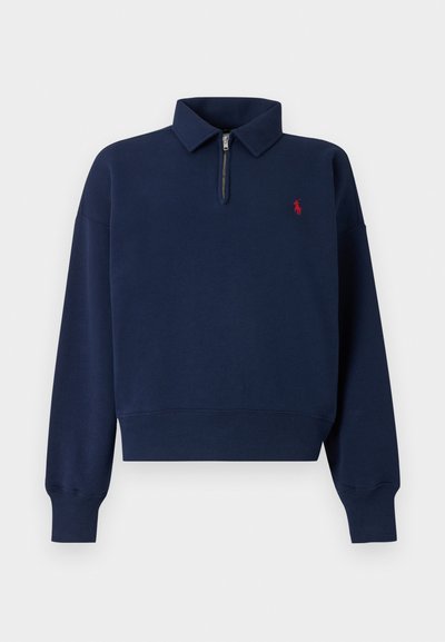 Tumesinine pullover kampsun poole lüpsuga ja soonikkoes varrukate äärtega. Vasakul rinnal on väike punane logo. Pehme puuvillane materjal, lõdvestunud lõige.
