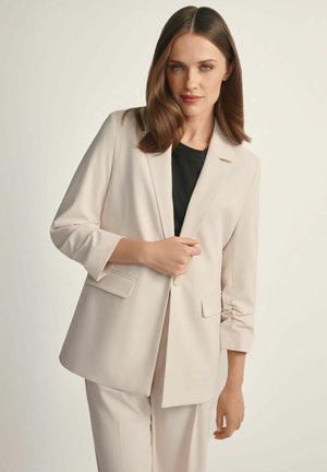 Kobieta o brązowych włosach do ramion, ubrana w beżową marynarkę i pasujące spodnie na czarnym topie, stojąca na jednolitym jasnym tle.