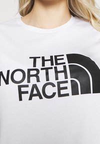 Bílé tričko s krátkými rukávy, na kterém se nachází výrazný černý text "THE NORTH FACE" a výrazný černý design polokruhového loga.