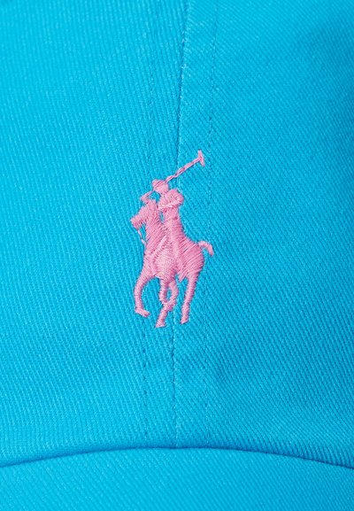 Mėlyna medvilninė kepurė, kurios priekyje yra rausvai siuvinėtas polo žaidėjo, jodinėjančio ant žirgo, logotipas. Audinys yra tekstūruotas, su matomomis siūlėmis.