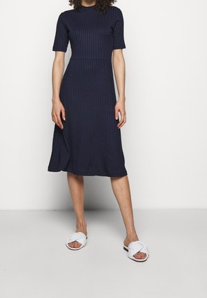 Personne portant une robe côtelée bleu marine longueur genou avec manches courtes, associée à des sandales blanches ouvertes à talons croisés, debout contre un mur blanc.