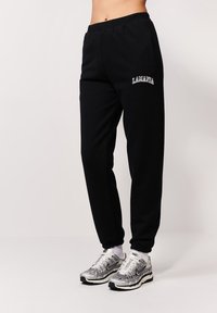 Czarne dresy z elastycznymi mankietami, z kieszeniami bocznymi i haftowanym logo "LAMANIA". W połączeniu z srebrnymi butami sportowymi i białymi skarpetkami.