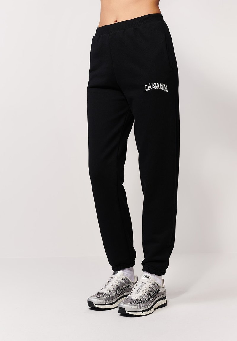 Czarne dresy z elastycznymi mankietami, z kieszeniami bocznymi i haftowanym logo "LAMANIA". W połączeniu z srebrnymi butami sportowymi i białymi skarpetkami.
