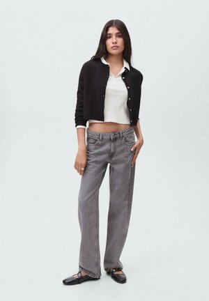 Kvinde iført sort cropped cardigan, hvid ribbet cropped polo, løse grå jeans og sorte Mary Jane flade sko, stående foran en ensfarvet baggrund.