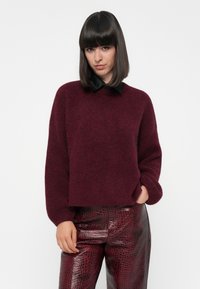 Burgunderfarbener Strickpullover mit überschnittenen Schultern, Rundhalsausschnitt und strukturiertem Material. Kombiniert mit roten Lacklederhosen im Reptilienmuster.