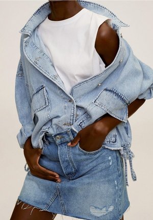 Persona lleva una chaqueta de mezclilla azul clara sobre una blusa sin mangas blanca y una falda de mezclilla desgastada a juego, con las manos en los bolsillos.