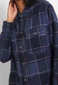 Camicia a quadri blu navy realizzata in morbido tessuto, con due tasche sul petto, chiusura a bottoni e linee viola chiaro nel motivo.