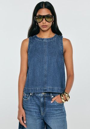 Kobieta nosząca oversize kwadratowe okulary przeciwsłoneczne, bezrękawnik z denimu, jeansy z wysokim stanem, złote kolczyki koła oraz masywne złote bransoletki z ręką w kieszeni.