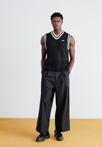 Black sleeveless sweater with white stripes, paired with wide black trousers and black shoes, set against a plain background.
