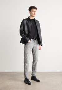 Jachetă din piele ecologică neagră cu buzunare frontale, purtată peste un tricou negru. Blugi ușor deteriorați gri deschis și cizme negre până la glezne completează look-ul.