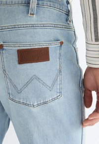 Lys blå Wrangler jeanslomme med brunt logopatch, kobbernitter, og en persons hånd hvilende ved siden av lommen med en stripete skjorte.