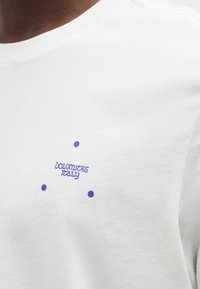 Κοντινό πλάνο λευκού πουκάμισου με μικρό μπλε κείμενο που γράφει "Dolomites Italy" περιτριγυρισμένο από τρεις μπλε τελείες στην περιοχή του στήθους.