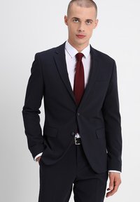 Dunkelblauer Anzug mit scharfen Revers, einreihigem Knopfverschluss und zwei vorderen Taschen. Kombiniert mit einem weißen Hemd und einer strukturierten burgunderroten Krawatte.