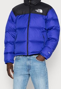 Mies, joka on pukeutunut sinimustaan The North Face -puffertakkiin, vaaleansinisiin farkkuihin, yksi käsi taskussa ja toinen rentona sivulla.