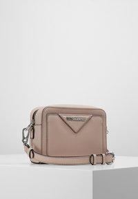 Pieni vaaleanbeige olkalaukku etutaskulla, hopea Karl Lagerfeld -logo, vetoketjullinen yläosa ja säädettävä hihna valkoisella pinnalla.