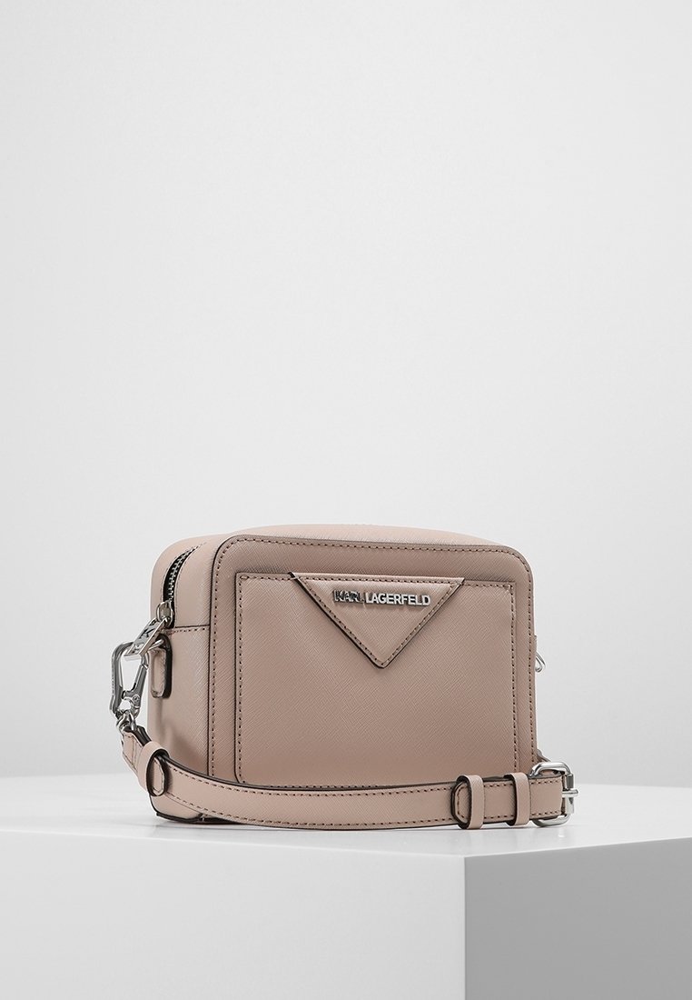 Pieni vaaleanbeige olkalaukku etutaskulla, hopea Karl Lagerfeld -logo, vetoketjullinen yläosa ja säädettävä hihna valkoisella pinnalla.