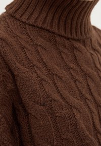Hnědý pletený svetr se žebrovaným vysokým límcem a texturovaným kabelovým vzorem, který zdůrazňuje jeho měkký a teplý materiál.