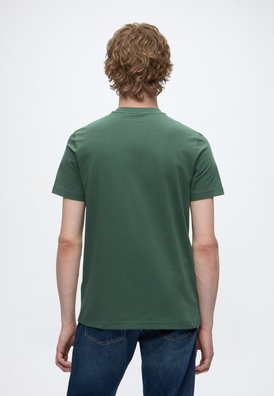 Personne aux cheveux bouclés châtain clair portant un t-shirt vert uni à manches courtes et un jean bleu, vue de dos sur fond blanc.