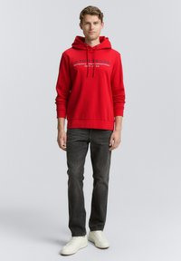 Un sweat à capuche rouge avec une poche kangourou à l'avant, orné d'un imprimé logo. Associé à un jean gris foncé et des baskets blanches.