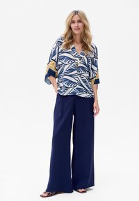 Luźna niebiesko-biała bluzka z nadrukiem liści, dekoltem w serek i szerokimi rękawami, w połączeniu z granatowymi spodniami o szerokich nogawkach i brązowymi sandałami.