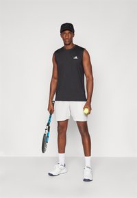 Camiseta atlética sin mangas negra, pantalones cortos gris claro, zapatillas de tenis blancas, sosteniendo una raqueta de tenis y una pelota, de pie contra un fondo liso.