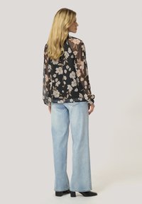 Blusa trasparente nera a fiori con maniche lunghe, decorata con fiori crema e pesca, abbinata a jeans a gamba larga azzurri e stivali neri con tacco.