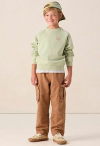 Sweat-shirt vert clair avec des poignets côtelés et un petit logo, associé à un pantalon cargo tan avec des poches latérales, et des chaussures beiges.
