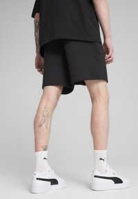 Shorts noirs à coupe décontractée, associés à des baskets blanches avec une bande noire. Tatouages remarquables visibles sur la jambe. Fond gris.