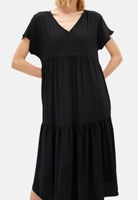 Žena v volném černém šatech s výstřihem do V a krátkým rukávem, s nabíranou sukní vrstvené, stojící s rukama v bok.