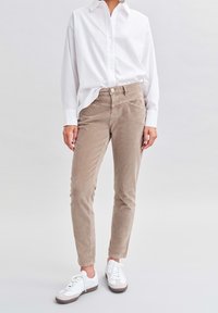 Pantalones de pana beige ajustados, con bolsillos frontales y cierre de botón, combinados con una camisa blanca de botones y zapatillas blancas.