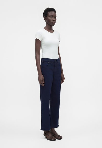 Γυναίκα με κοντά κατσαρά μαλλιά, που φοράει λευκή εφαρμοστή μπλούζα με κοντά μανίκια, σκούρο μπλε παντελόνι με ίσια πόδια και μαύρα σανδάλια, στέκεται μπροστά σε ένα μονόχρωμο φόντο.