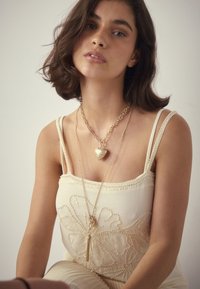 Young woman with short dark hair wears a cream dress with beaded floral patterns and two gold necklaces, one with a heart pendant.