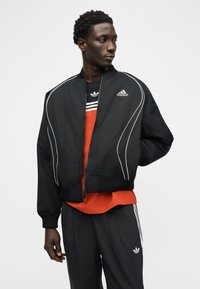 Νεαρός άνδρας φορώντας μαύρο αθλητικό μπουφάν και παντελόνι Adidas με κόκκινο πουκάμισο από κάτω, στέκεται με το ένα χέρι πίσω από την πλάτη, κοιτάζοντας προς το πλάι.