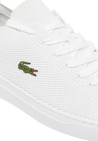 Biały but sportowy z teksturowaną siateczkową cholewką, płaskimi białymi sznurówkami i zielonym logo krokodyla po boku. Gumowa podeszwa z minimalnym bieżnikiem.