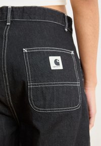 Pantaloni in denim neri con cuciture bianche, dotati di una tasca posteriore e di una patch quadrata con il logo. Il materiale appare robusto e ha una texture liscia.