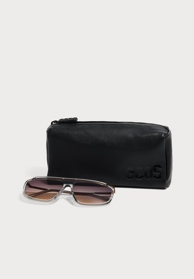 Lunettes de soleil rectangulaires à dégradé marron avec montures transparentes, posées devant une pochette zippée en cuir noir avec lettres en relief.
