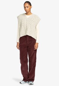 Light gray cable-knit sweater vest with a v-neck, paired with burgundy corduroy pants. Model stands on a plain background. Silver sneakers.