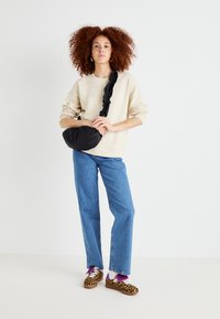 Schwarze gepolsterte Umhängetasche mit einem gerafften Schulterriemen, getragen über einem cremefarbenen Sweatshirt, kombiniert mit blauen geraden Jeans und Schuhen im Leopardenmuster.