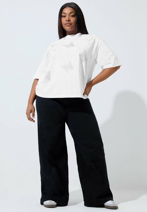 Kvinde står med hånden på hoften iført hvid T-shirt med sommerfuglprint, sorte bukser med vide ben og hvide sneakers mod en ensfarvet baggrund.