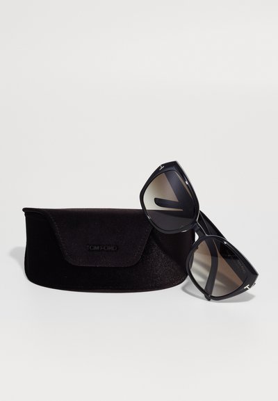 Svarta Tom Ford-solarier med gradientlinser, placerade i en vinkel bredvid ett svart sammetetui. Etuiet har en lockstängning och en logotyp.