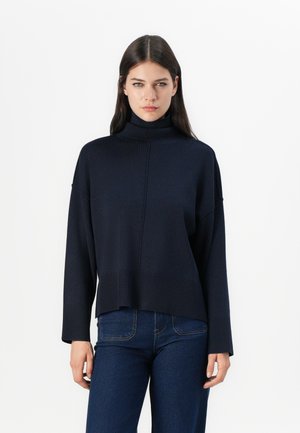 Dunkelblauer Rollkragenpullover mit einem lässigen Schnitt, geripptem Saum und Nahtdetails. Getragen mit dunklen Jeans. Weiche Strickstruktur.