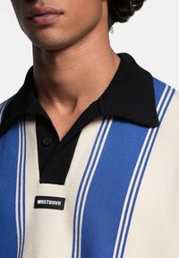 Polo skjorte med sort krave, prydet med lodrette blå og cremefarvede striber, blød tekstur og en sort label med "WRSTBHVR" logo ved knapstolpen.