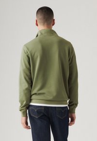 Olijfgroene sweatshirt met een hoge kraag, lange mouwen en een zachte textuur. Gepaard met donkerblauwe jeans, waarbij de details van de achterkant en de mouwen zichtbaar zijn.
