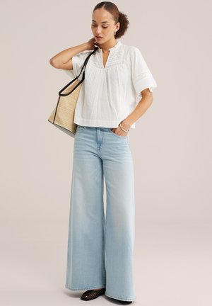 Donna che indossa una camicetta bianca ricamata, jeans azzurri con gamba larga, scarpe nere e che porta una borsa tote intrecciata color cuoio con rifiniture nere.