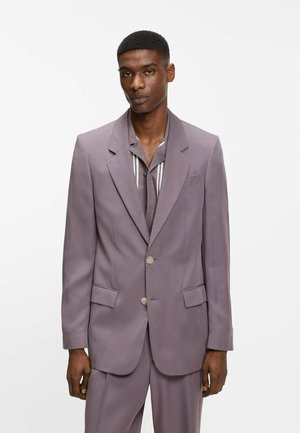 Mand iført en lilla/dråbe farvet jakkesæt med to knapper fastgjort, over en lilla skjorte med lodrette hvide striber, stående mod en ensfarvet hvid baggrund.