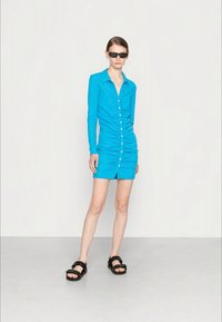 Vestido longo de mangas compridas em turquesa com detalhes franzidos e botões brancos, combinado com sandálias pretas. Modelo usa óculos de sol escuros.