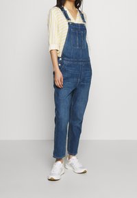 Jeansoverall med justerbara remmar, frampocket och smalnade ben, bärs över en randig gul och vit skjorta, ihop med vita sneakers.