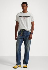 Λευκό κοντομάνικο μπλουζάκι με μαύρη και κόκκινη εκτύπωση "POLO SPORT", συνδυασμένο με μπλε τζιν παντελόνι και κίτρινα αθλητικά παπούτσια με μπλε λεπτομέρειες.